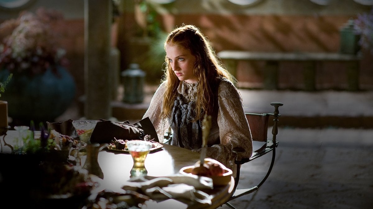 Sophie Turner as Sansa Stark sits in tense contemplation during a pivotal Game of Thrones scene, dressed in an intricately embroidered, high-neck gown that reflects her nobility and growing inner strength
