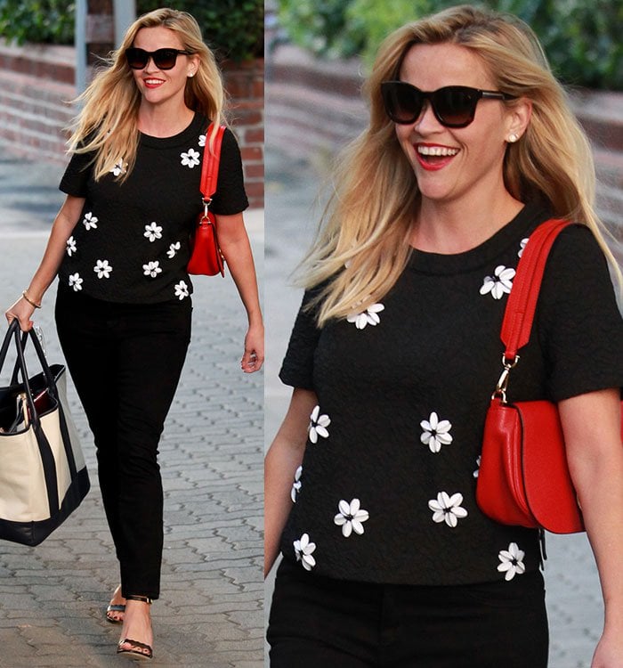 Reese Witherspoon steps out in a chic black-and-white floral top paired with sleek black pants, a bold red handbag, and classic heels