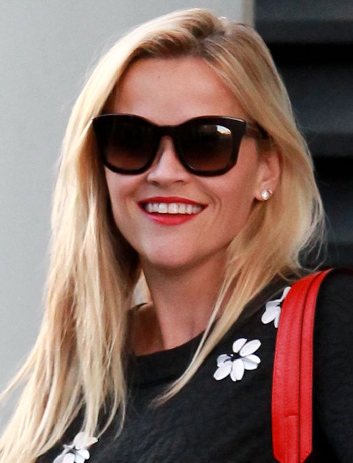 Reese Witherspoon flashes a bright smile, accessorizing her casual look with oversized sunglasses and a pop of red lipstick
