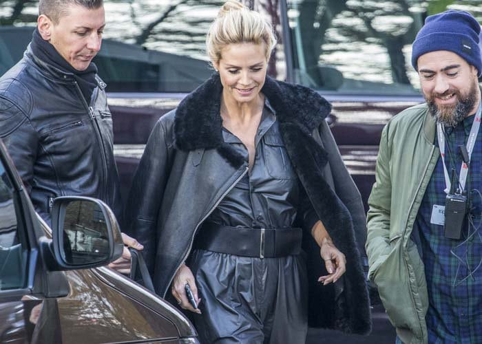Heidi Klum, with her hair in an elegant updo, is seen in a black leather jacket with a fur collar, a dark button-up shirt, matching pants cinched with a wide belt, and is accompanied by two men