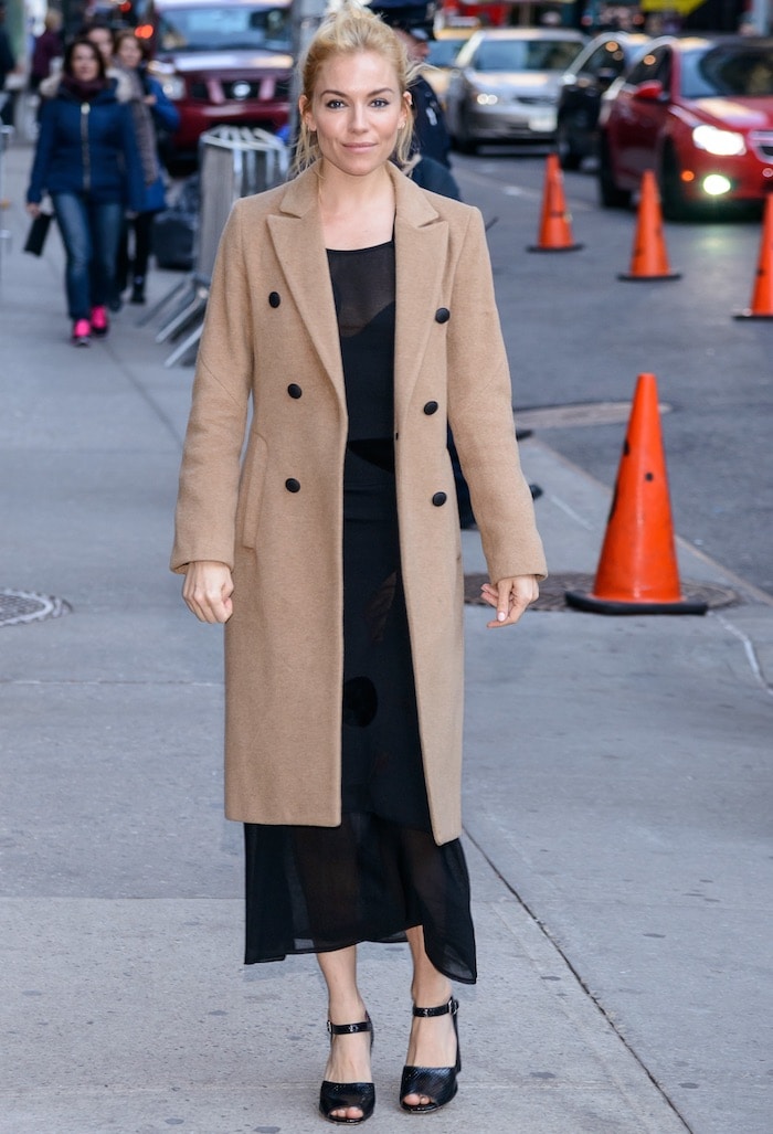 Sienna Miller layers a classic camel coat over a sheer black Roland Mouret dress, finishing the look with bold snakeskin-textured block-heeled sandals in New York City