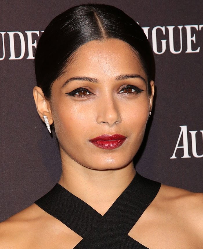 Freida Pinto finishes her Audemars Piguet look with a sleek center-parted bun, winged eyeliner, deep red lipstick, and diamond drop earrings
