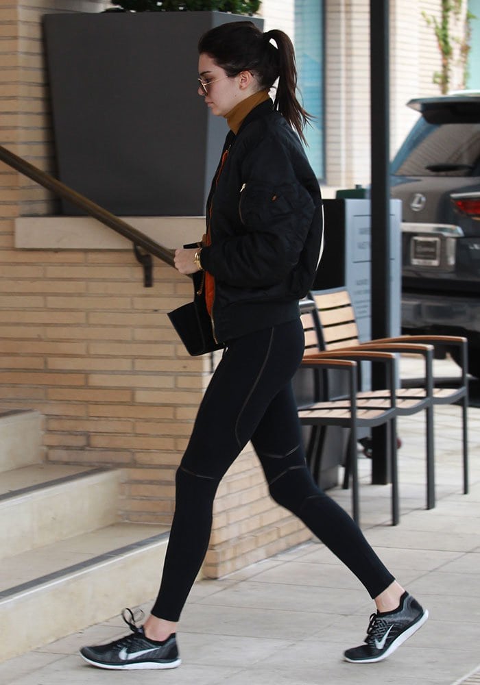 Kendall Jenner opts for a sporty-chic look with a black bomber jacket, toffee-colored cropped turtleneck, and black Nike Flyknit sneakers