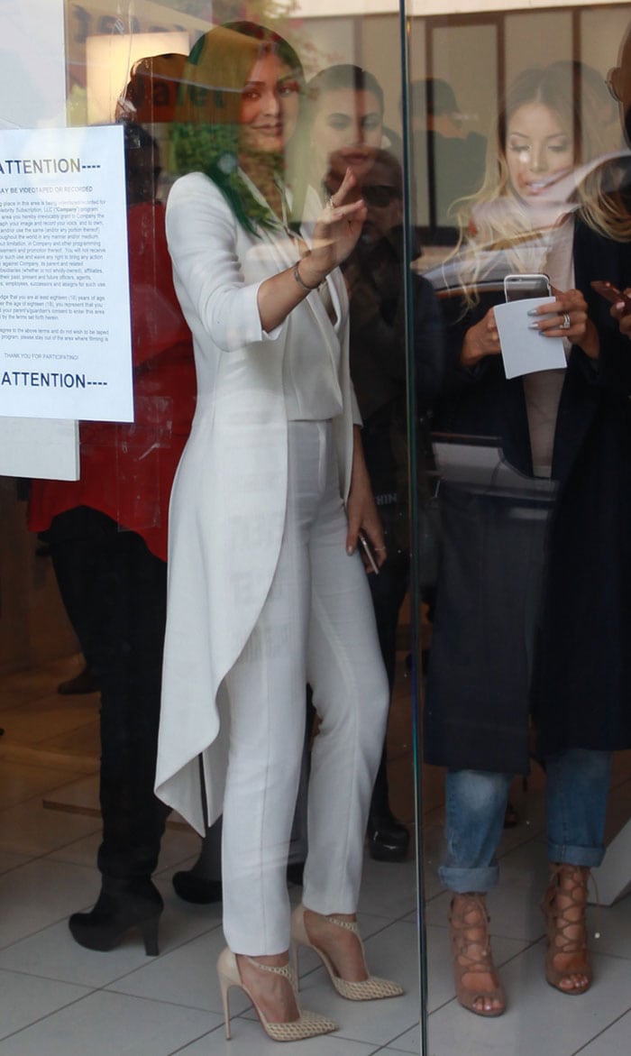 Pausing behind glass doors, Kylie flashes a peace sign while her structured white tailoring and sheer pumps maintain a polished, minimalist silhouette