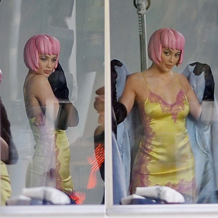 Seen through a studio window, Kylie Jenner strikes more poses in a molded pink wig and a yellow slip dress with pink lace trim