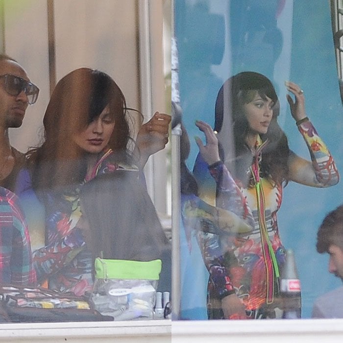 Through a window, Kylie Jenner adjusts her black wig while posing in a vibrant zip dress with a distinctive birds-and-flowers print