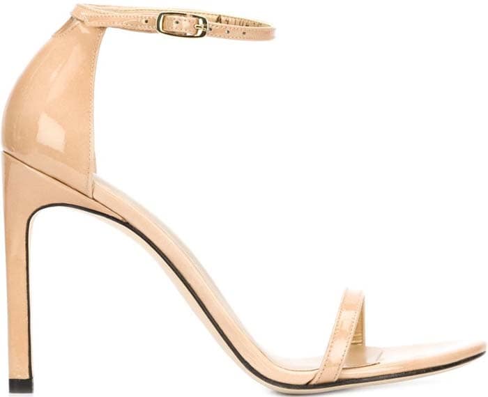 The nude patent Stuart Weitzman “Nudistsong” sandal distills evening dressing to its essentials, with a slim toe strap, delicate ankle wrap, and softly curved stiletto that lengthens the leg line without competing for attention