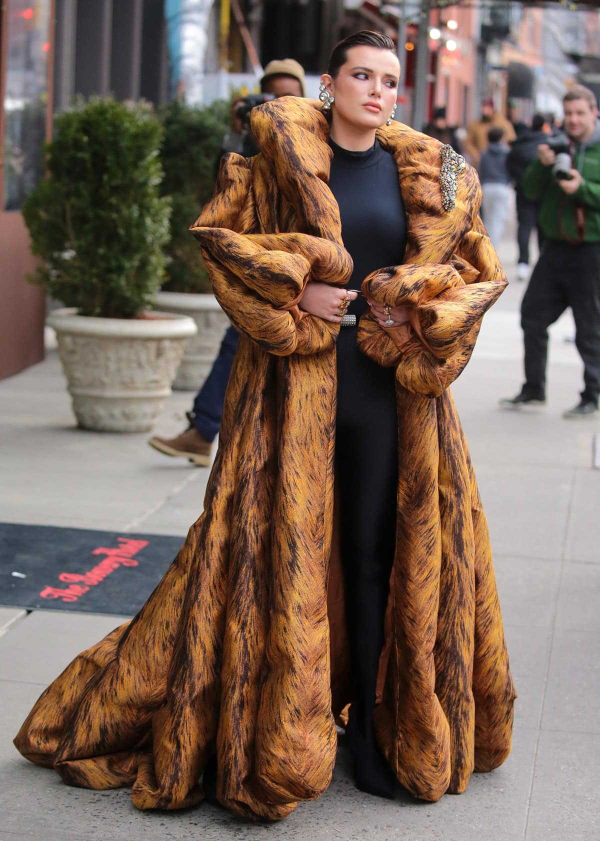 Bella Thorne embraced the Mob Wife trend, wearing a striking orange and black puffy coat with a chapel train, accessorized with a large brooch and crystal-encrusted earrings, along with a sleek black sweater, leggings, and booties at New York Fashion Week
