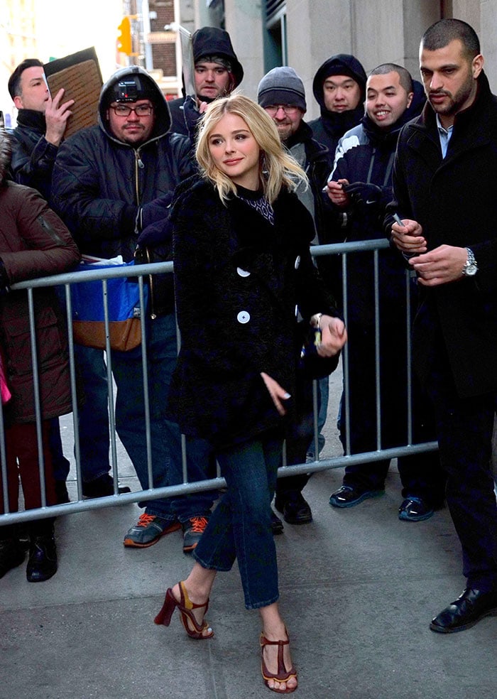 Chloë Grace Moretz wears a black coat over a high-neck top and a pair of three-quarter length jeans
