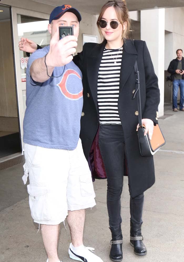 Dakota Johnson pauses to take a fan photo at the airport, wearing a black double-breasted coat over a striped tee and skinny jeans, finished with studded ankle boots and round sunglasses that keep the moment relaxed yet polished