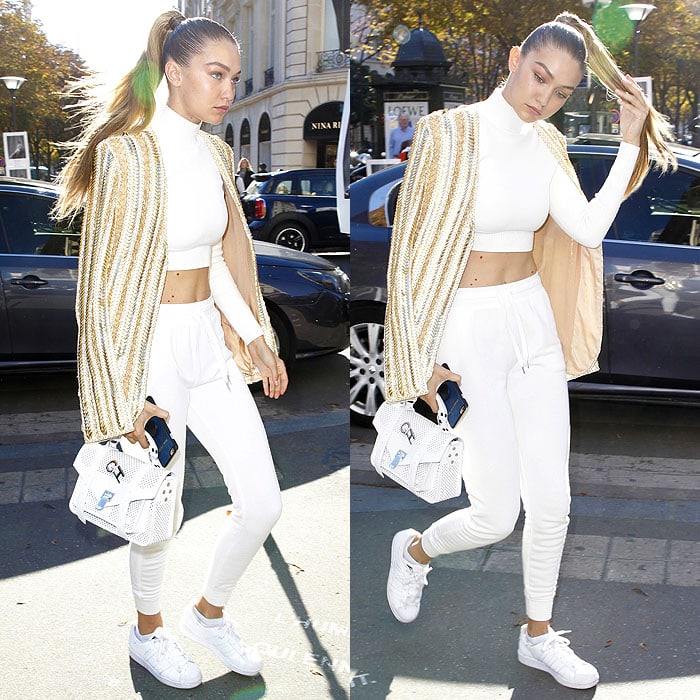 Gigi Hadid brings white sneakers onto the fashion week circuit, grounding a cropped all-white look and embellished jacket with clean trainers while arriving at the Balmain show