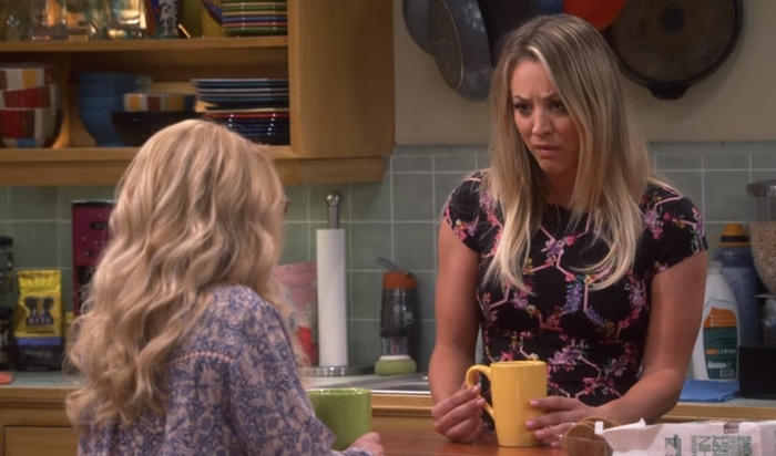 Kaley Cuoco as Penny shares a tense kitchen conversation with Melissa Rauch’s Bernadette, clutching a yellow mug while wearing a fitted molecule-print top