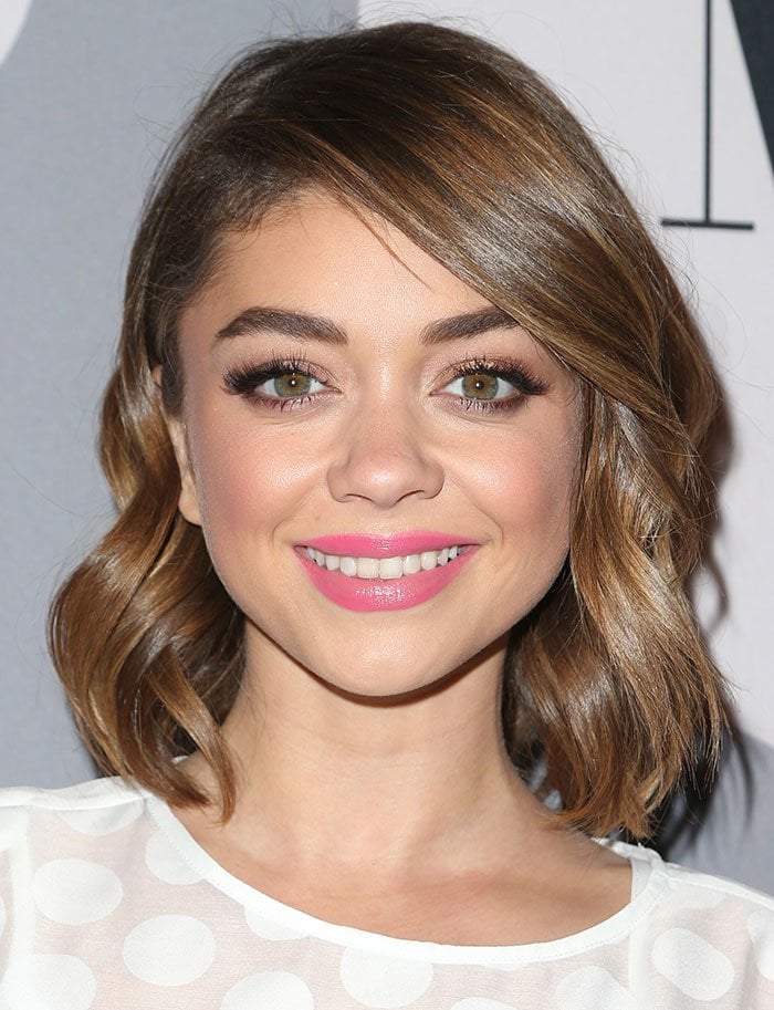 Sarah Hyland complements her sheer white polka-dot ensemble with softly waved hair, fluttery lashes, and bright pink lipstick