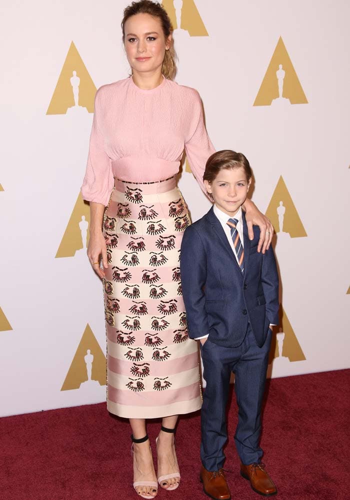Brie Larson, wearing an Emilia Wickstead pink puff-sleeve top, embroidered midi skirt, and Christian Louboutin PVC-and-suede ankle-strap sandals, poses with her Room co-star Jacob Tremblay in a sharp blue suit
