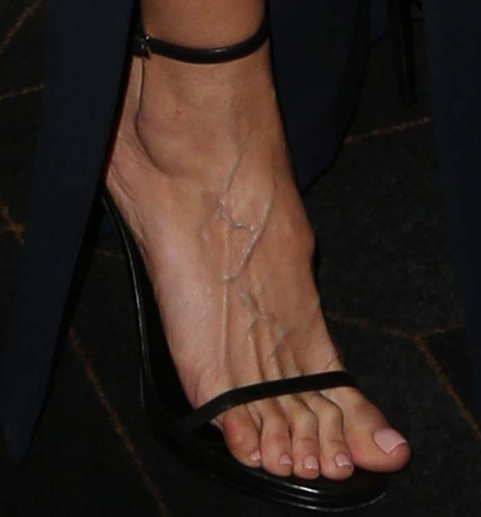 A close-up shows Heidi Klum's foot in black Stuart Weitzman "Nudist" sandals, highlighting their minimalist two-strap design