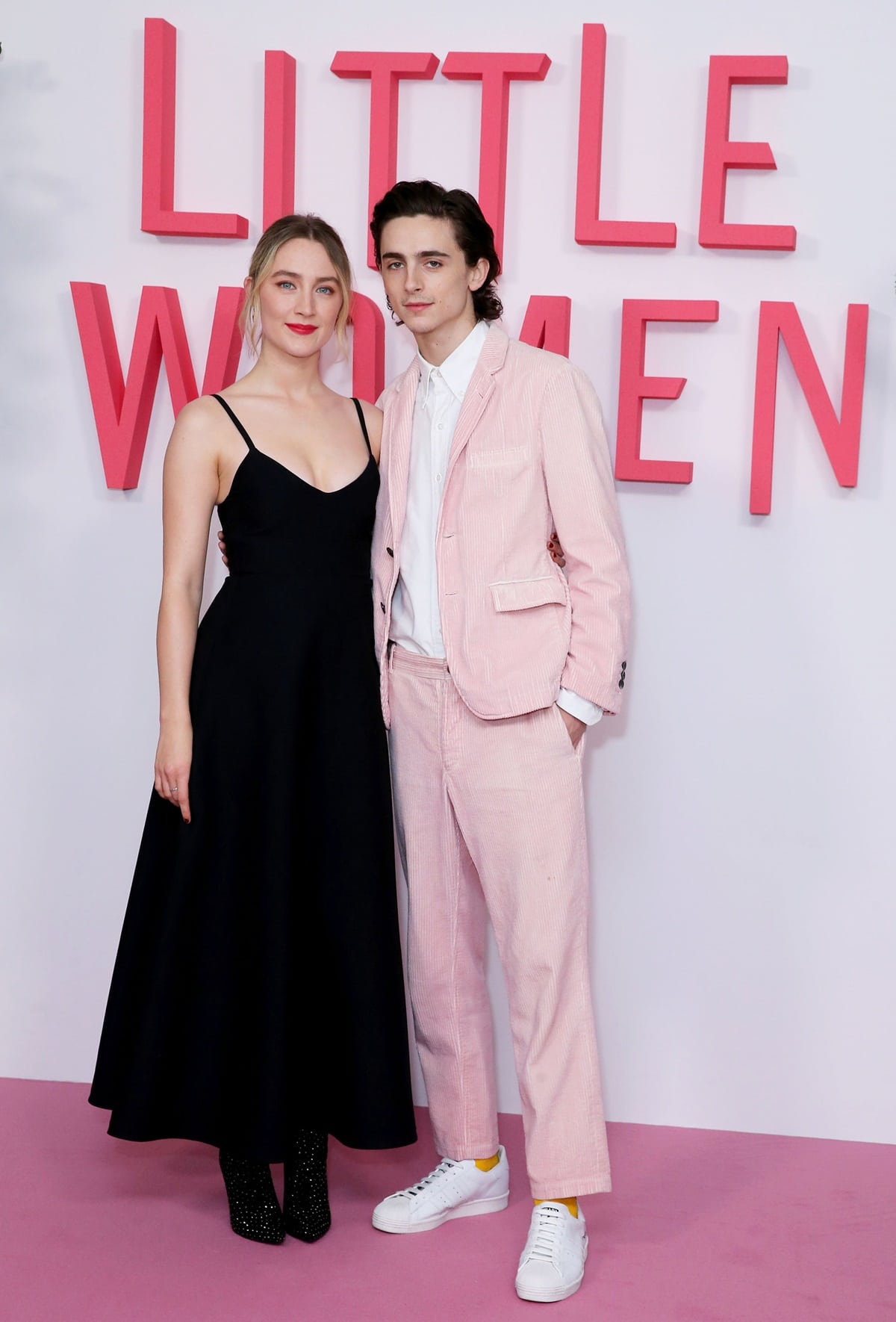 Saoirse Ronan paired a black Valentino Spring 2020 midi dress with glittery Nina boots and Theodora Warre earrings, while Timothée Chalamet contrasted in a pastel pink Thom Browne corduroy suit with yellow socks and white sneakers at the Little Women London premiere on December 16, 2019