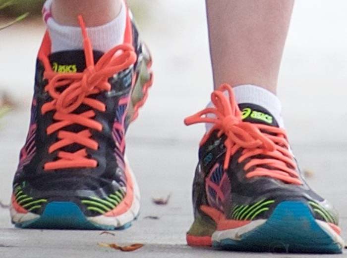 A close-up reveals Anne Hathaway’s colorful Asics Gel-Kinsei trainers, with bright coral laces and cushioned soles designed for stability and long-distance comfort on the trail
