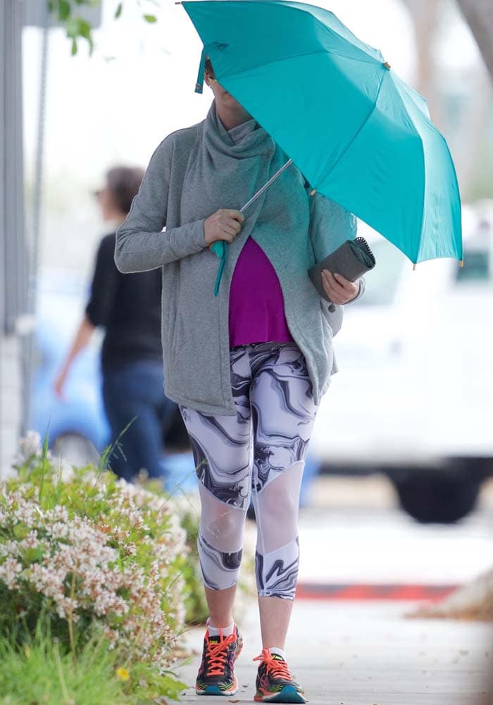 Anne Hathaway keeps things low-key while heading out in Los Angeles on March 15, 2016, layering a gray zip-front jacket over a bright pink top, Chillbywill Love crop leggings, and her go-to Asics Gel-Kinsei 6 sneakers, finishing the look with a teal umbrella for off-duty practicality