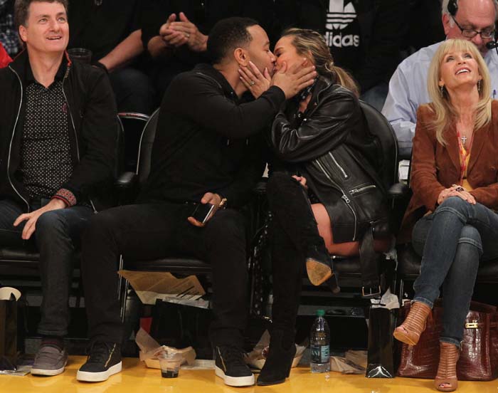 John Legend and Chrissy Teigen share a kiss for the kiss cam at a Los Angeles Lakers game, with Teigen wearing Chloé suede and textured-leather over-the-knee boots styled with an Acne leather jacket
