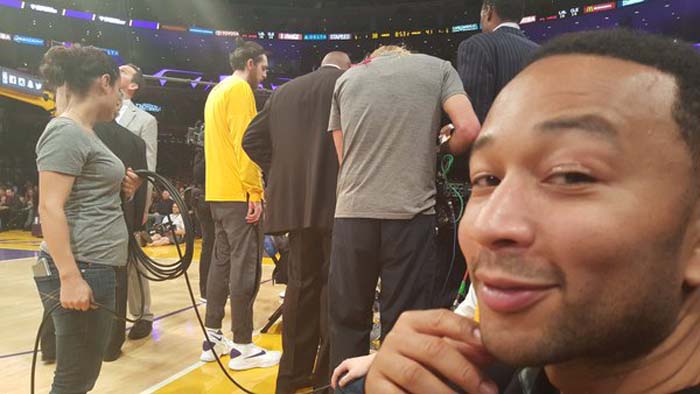 John Legend sitting courtside at Staples Center wearing a black jacket during a Lakers vs Cavaliers game