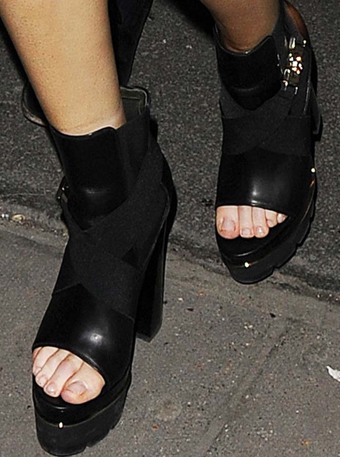 A close-up reveals the sculptural open-toe Versace platforms, featuring thick ankle cuffs, crisscross leather panels, and a bold stacked heel