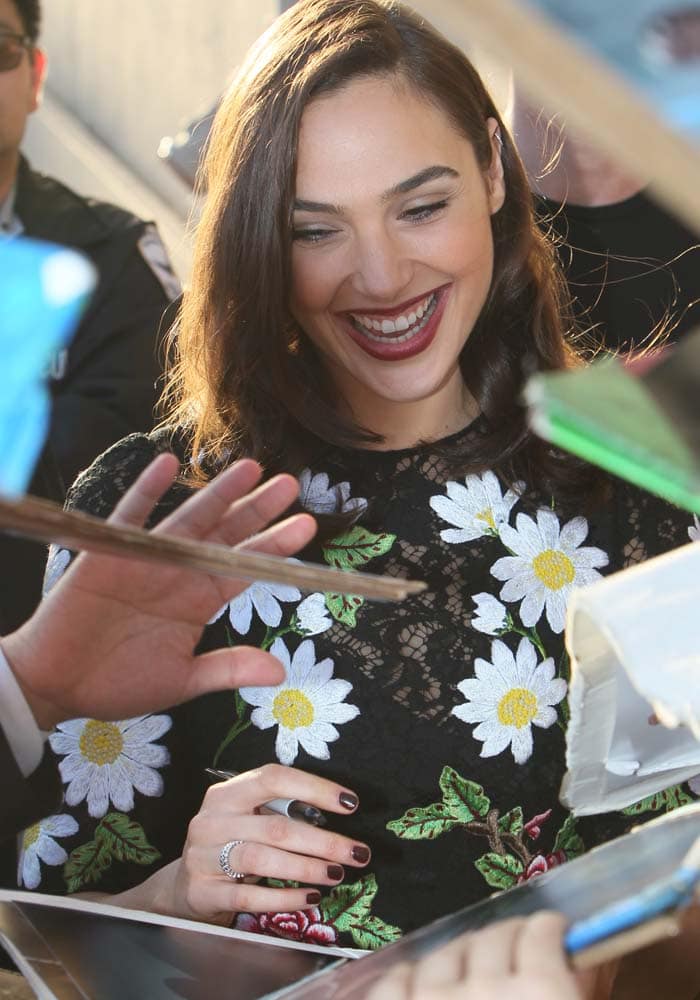 Gal Gadot smiles warmly as she signs autographs for fans, showing off the floral embroidery of her lace Dolce Gabbana mini dress