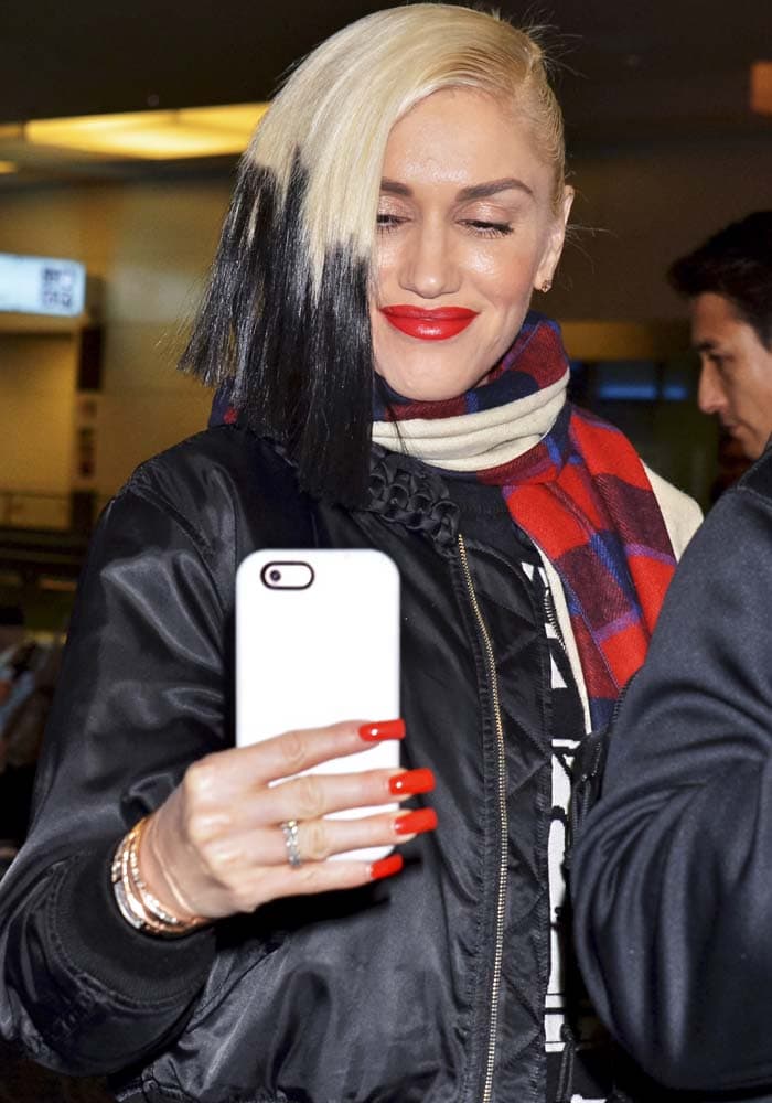 Gwen Stefani checks her phone at Tokyo’s Haneda Airport in a Moschino sweater layered under a Noir Kei Ninomiya jacket, finishing the look with a cozy Donni Charm scarf and her signature bold red lip