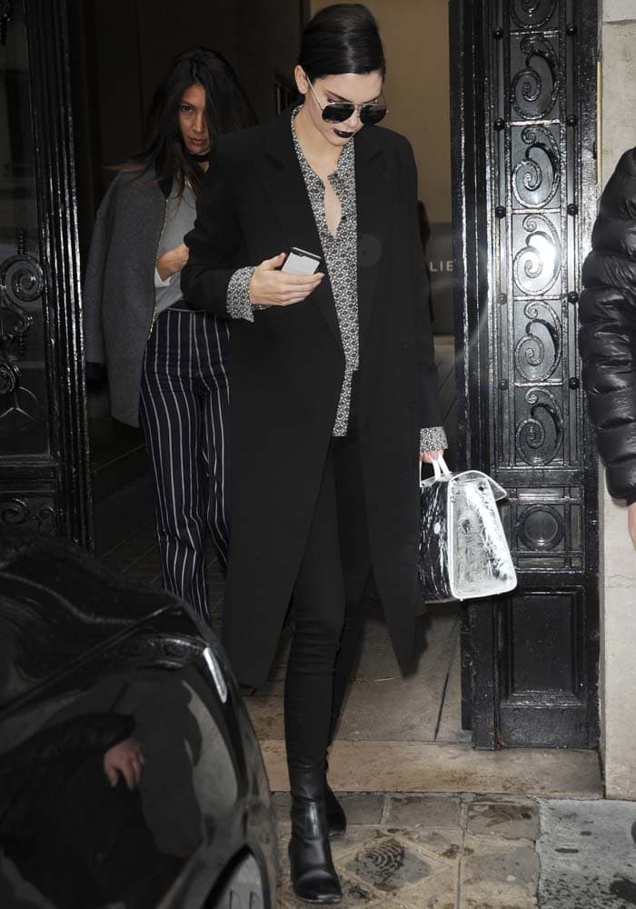 Kendall Jenner steps out in Paris in a sleek black coat, patterned blouse, black skinny jeans, and her go-to Sandro 'Amele' ankle boots, accessorized with dark sunglasses and a structured top-handle bag