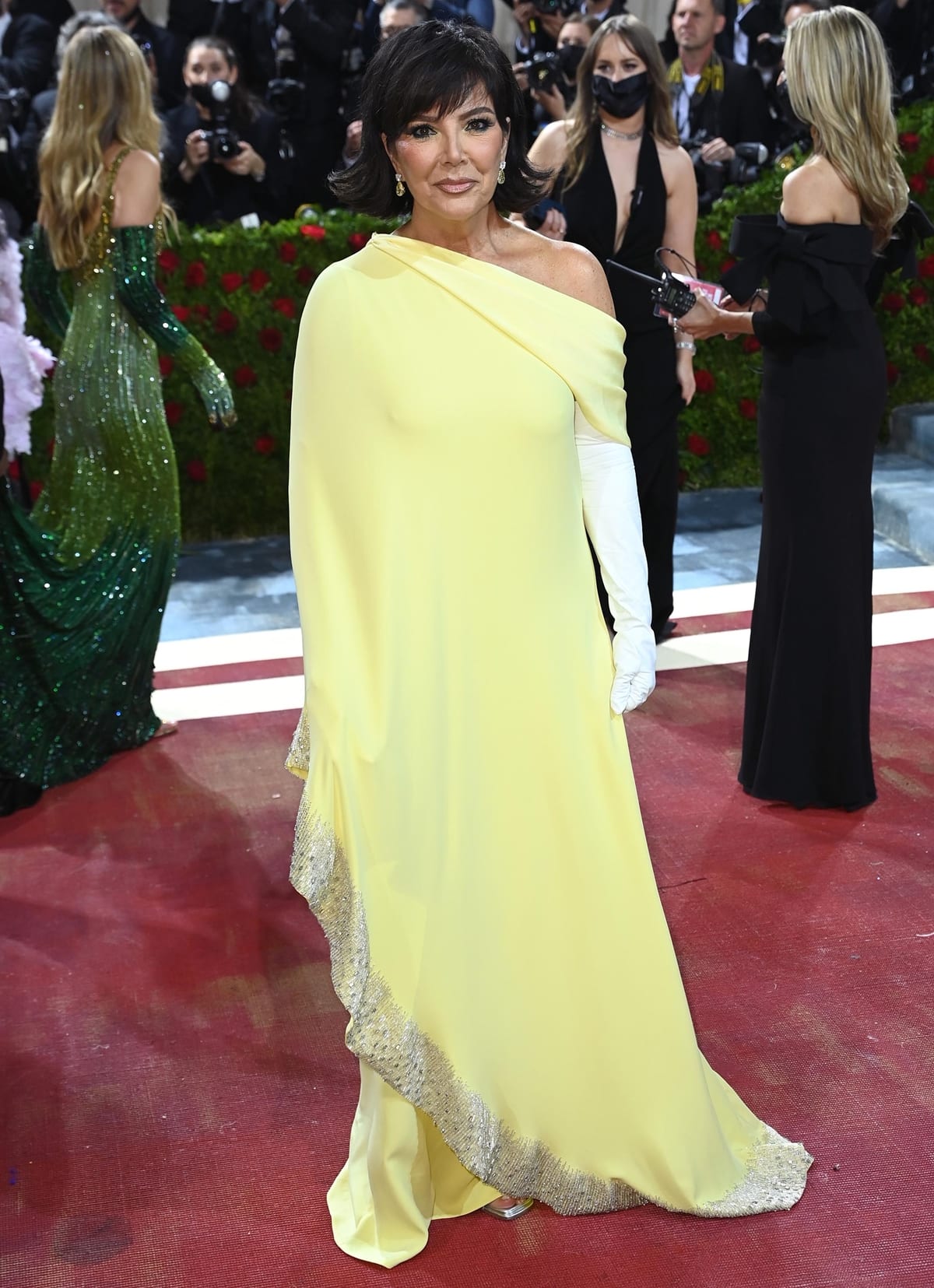 Kris Jenner attends the 2022 Met Gala in a flowing yellow silk Oscar de la Renta gown with white gloves and metallic platform heels peeking through the hem, blending Old Hollywood elegance with a modern gilded touch