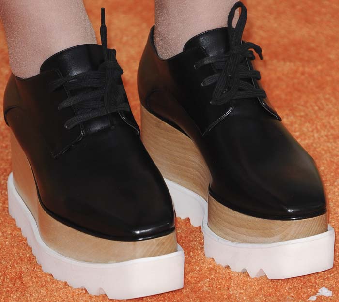 A close-up highlights Meghan Trainor’s Stella McCartney Elyse platform sneakers, showcasing the sleek black Derby-style upper set atop the signature saw-edged white sole and sculptural wooden wedge that define the cult vegan design