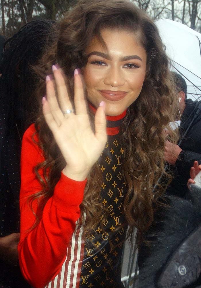 Zendaya waves in a Louis Vuitton monogram leather dress with bold red sleeves, wearing her hair in voluminous curls at Paris Fashion Week 2016