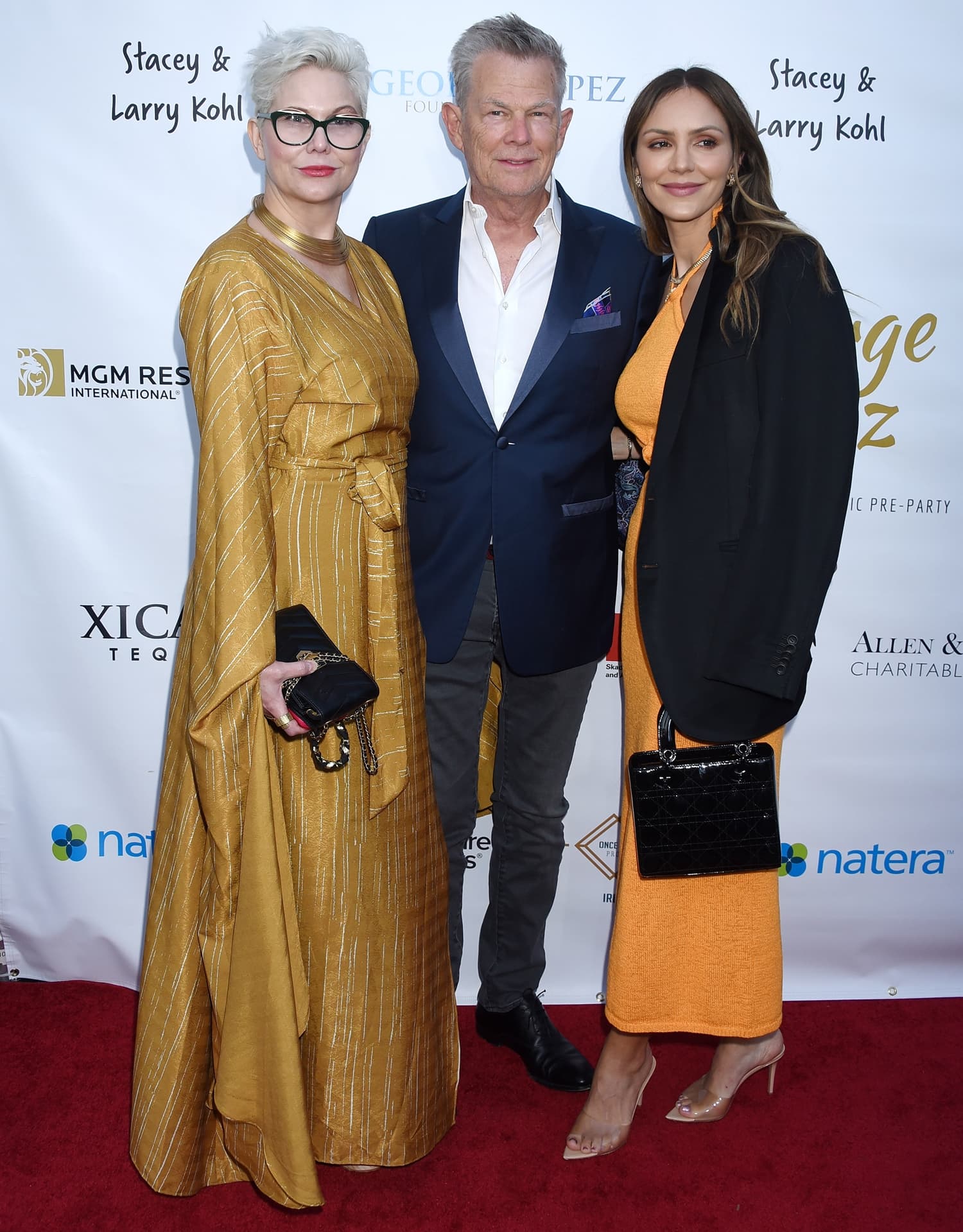 Katharine McPhee elongates her frame in barely-there clear heels, styling them with an orange midi dress, oversized black blazer, and structured patent handbag alongside David Foster and Amy S. Foster