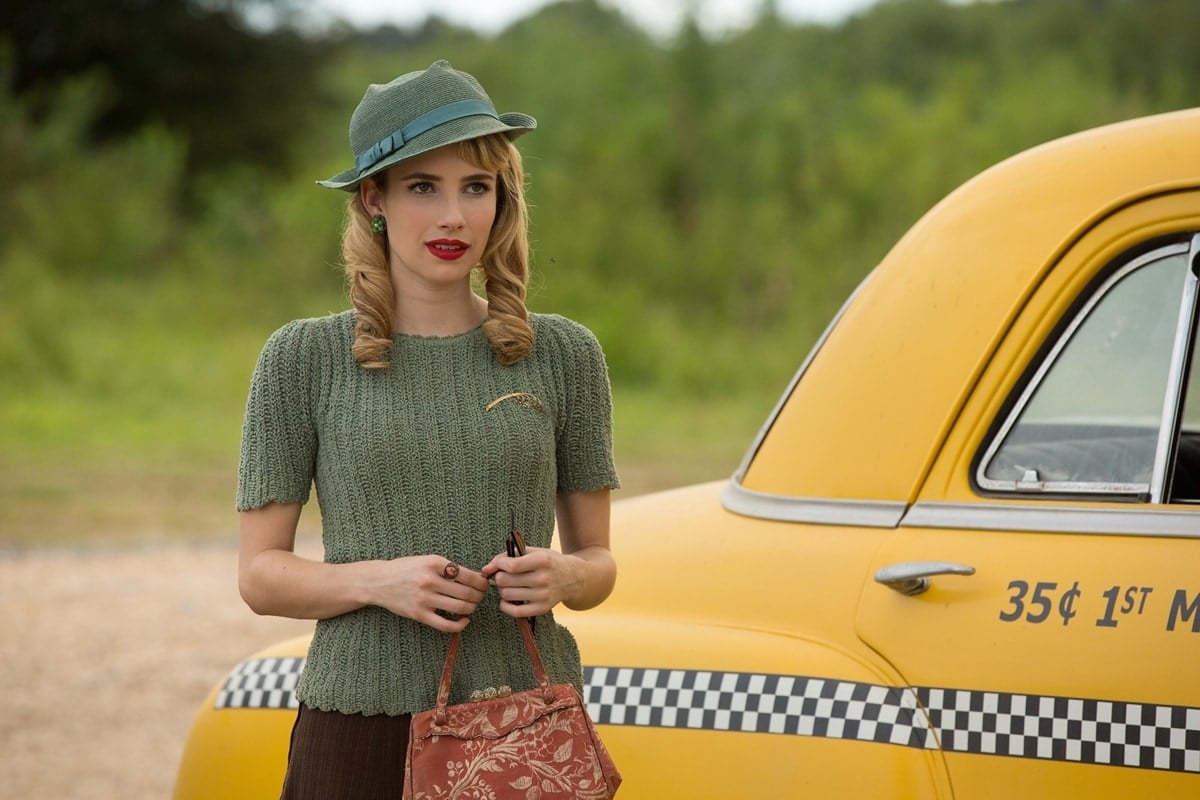Emma Roberts channels vintage elegance as fortune teller Maggie Esmerelda in American Horror Story: Freak Show—styled in a textured sage knit top, retro hat with ribbon trim, and a tapestry handbag that captures the show’s 1950s-inspired aesthetic
