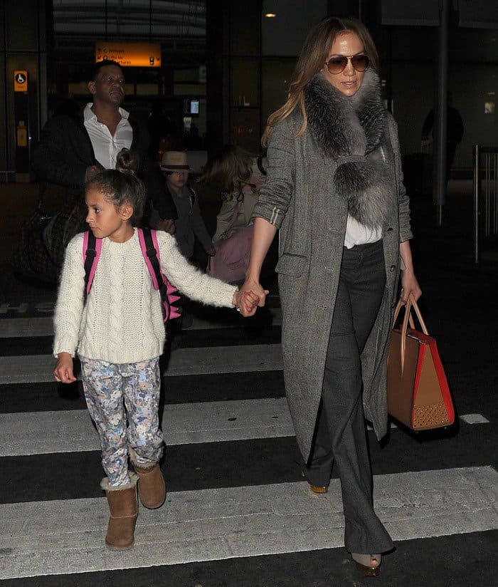Jennifer Lopez arrives at Heathrow Airport in London with her daughter, who pairs a cozy cream cable-knit sweater, floral joggers, and classic chestnut UGG boots for a practical travel look that contrasts with her mother’s polished tailoring