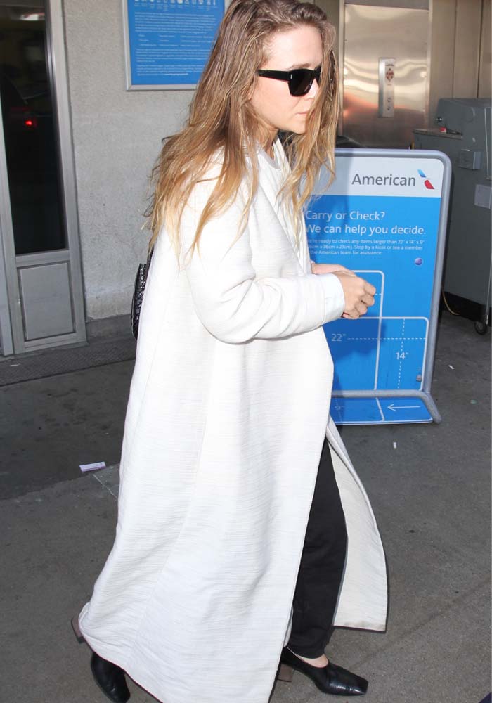 Mary-Kate Olsen keeps it effortlessly chic at LAX on April 2, 2016, wearing a full-length ivory coat over black trousers, sleek black mules, and Oliver Peoples × The Row 71st Street sunglasses, finishing her look with The Row’s alligator backpack and a Cartier engagement ring