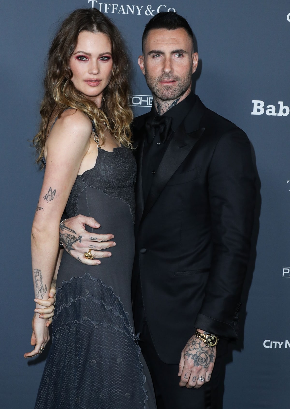 Behati Prinsloo stuns in a dark lace-trimmed gown paired with black platform heels while posing with Adam Levine, both showcasing their tattoos at the 2021 Baby2Baby Gala in West Hollywood