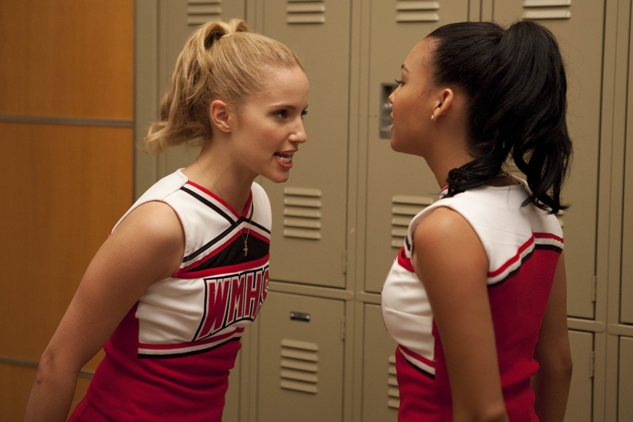 Naya Rivera as Santana Lopez and Dianna Agron as Quinn Fabray wear their signature WMHS cheerleading uniforms in a tense locker room scene from the hit series Glee, highlighting the drama that often unfolded behind the pom-poms