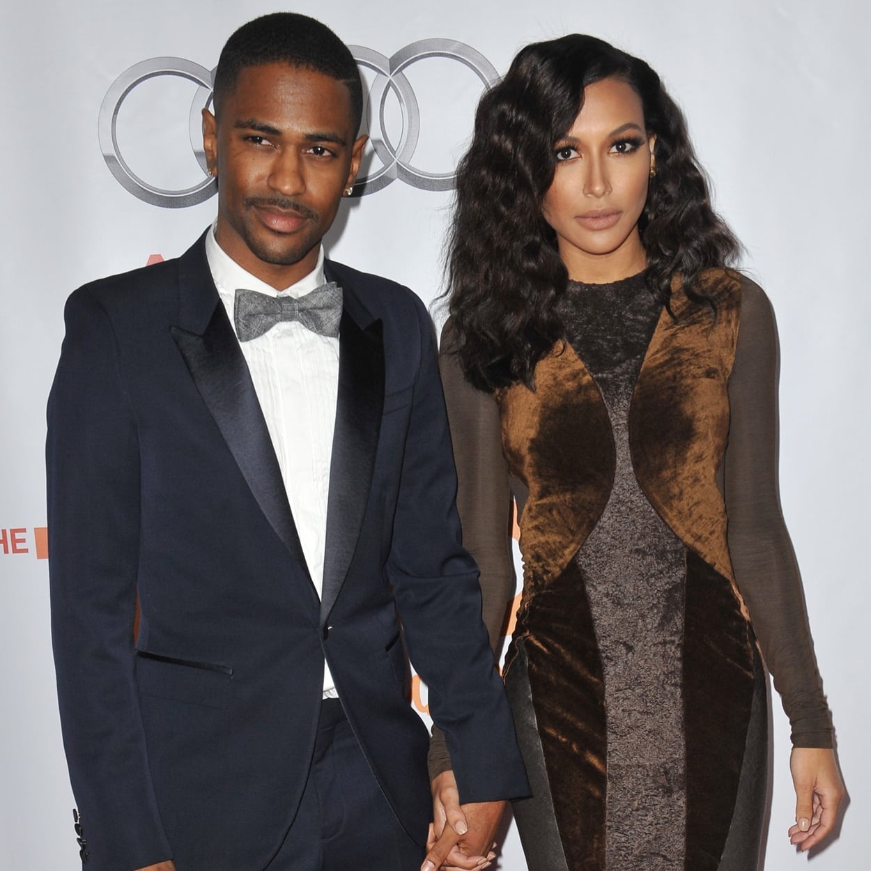 Naya Rivera stuns in a form-fitting, velvet-paneled dress with sheer long sleeves, while Big Sean complements her look in a classic navy tuxedo and silver bow tie at a red carpet event