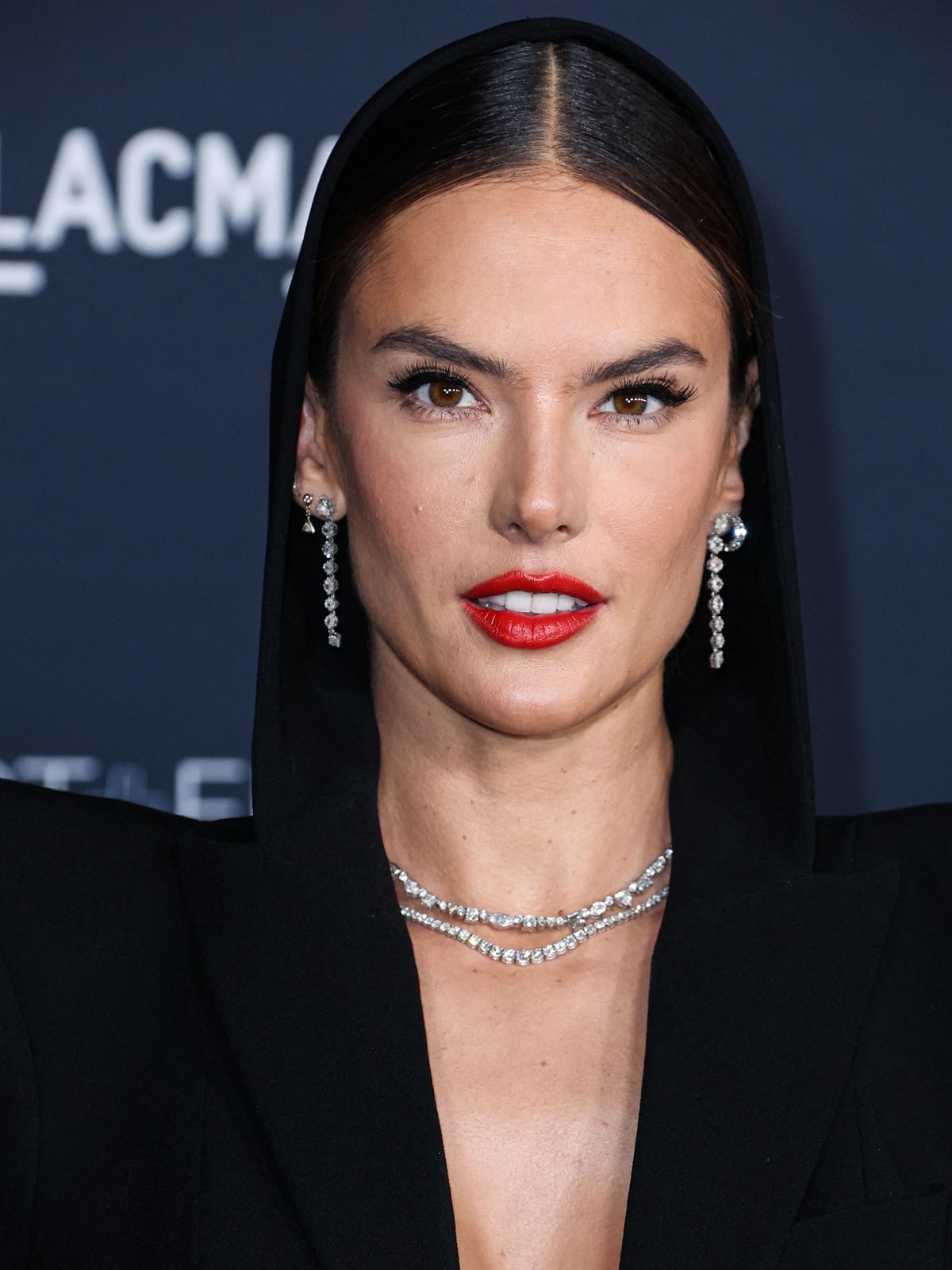 Alessandra Ambrosio stuns in a sleek black hooded Jean Paul Gaultier Couture outfit, accentuated by dazzling drop earrings and a bold red lip at the 2022 LACMA Art + Film Gala