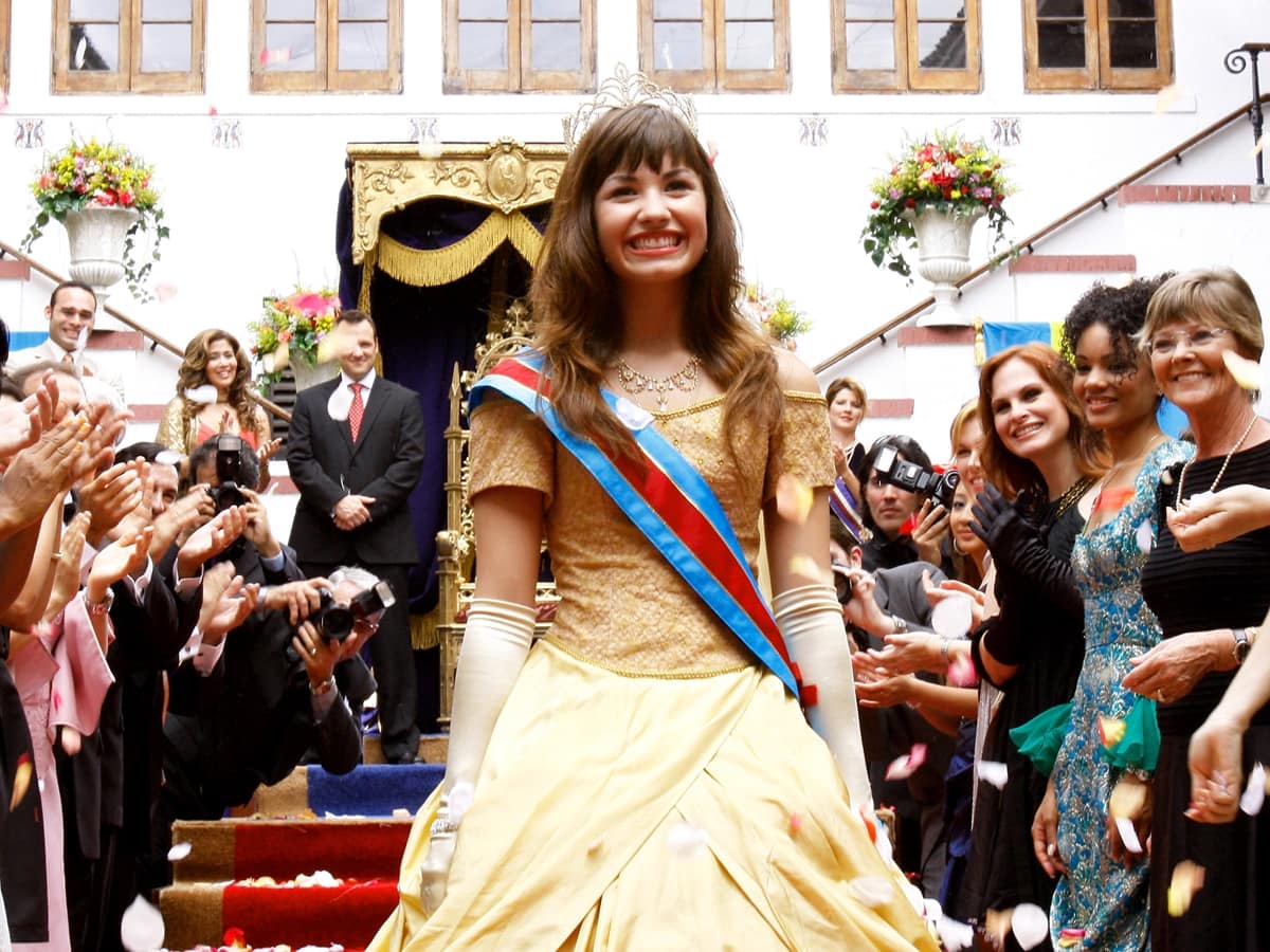 Demi Lovato, just 15 at the time, transforms into royalty in a golden off-the-shoulder gown with satin gloves and a sash as Princess Rosalinda María Montoya Fioré in Princess Protection Program