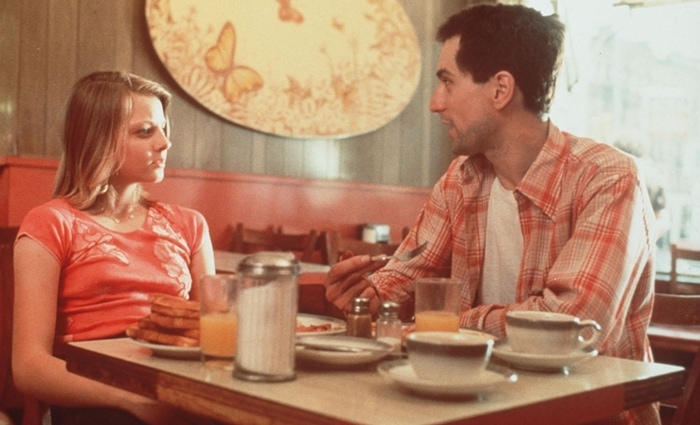 Jodie Foster’s Iris sits across from Robert De Niro’s Travis Bickle in Taxi Driver, contrasting her casual fitted T-shirt with his plaid shirt and everyday workwear
