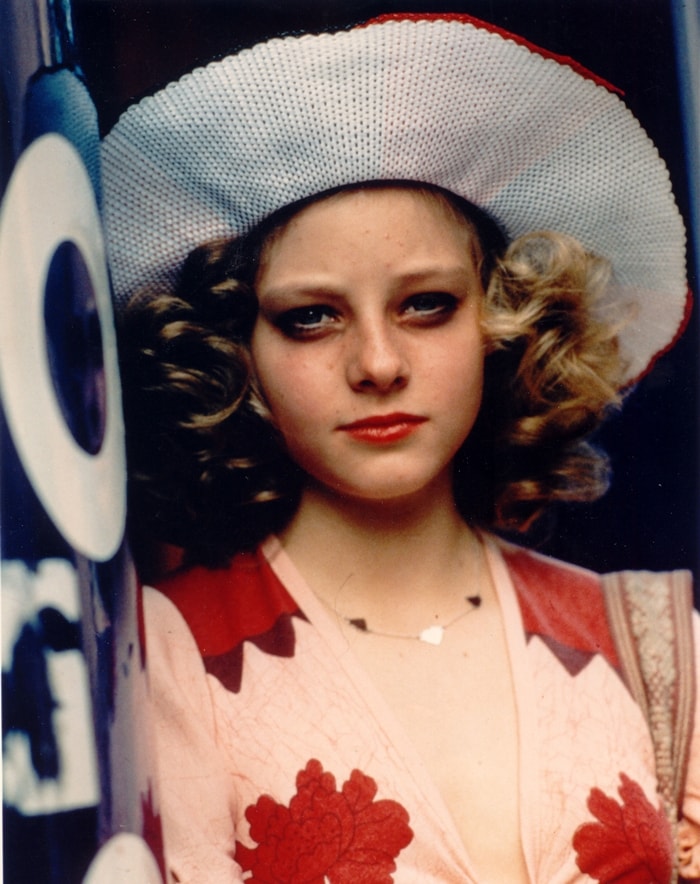 Jodie Foster as Iris in Taxi Driver, wearing a wide-brimmed hat, curled pigtails, heavy eye makeup, and the character’s deliberately adult-styled 1970s wardrobe