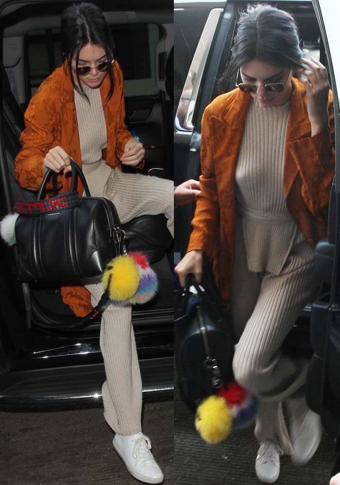 Kendall Jenner steps out in ribbed Elle Sasson separates layered beneath a burnt-orange Etro coat, accessorizing with a black Givenchy “Lucrezia” tote adorned with playful Fendi smiley pom-pom keychains while finishing the relaxed airport look with crisp white Kenneth Cole “Kam” sneakers
