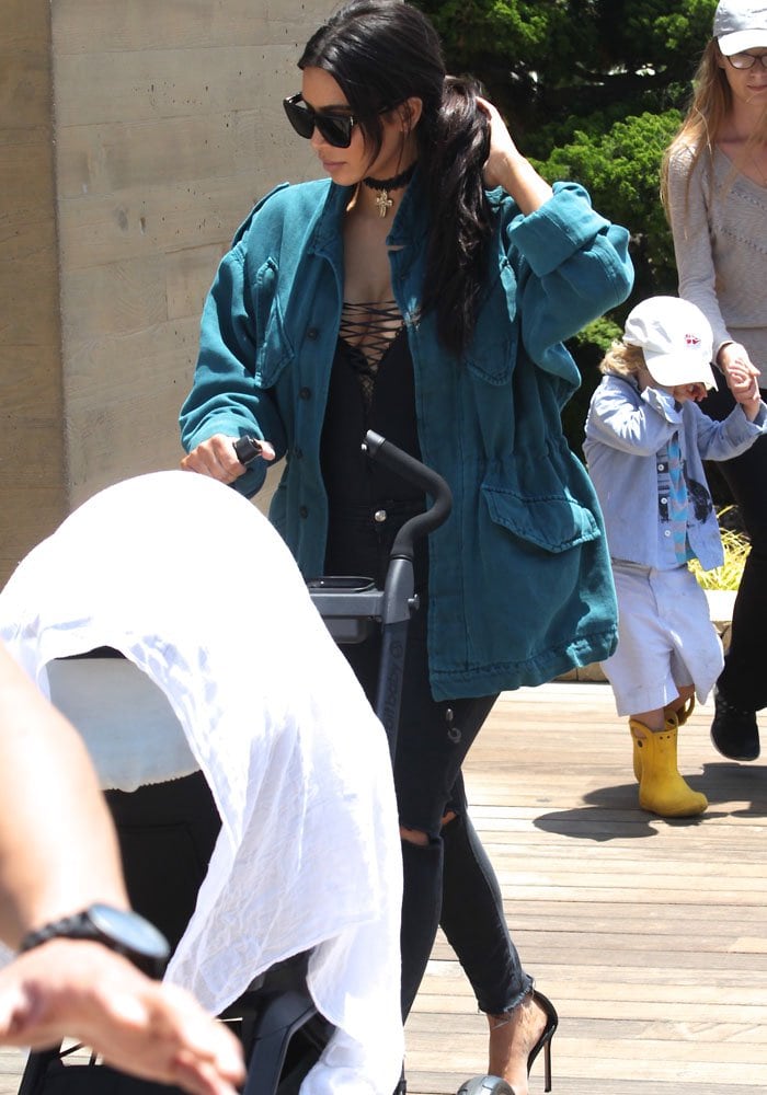 Kim Kardashian keeps her daytime look polished yet practical in Givenchy Gv 7002/S sunglasses, a lace-up black bodysuit layered under a relaxed jacket, J Brand Photo Ready cropped jeans in Mercy, and Manolo Blahnik suede “Chaos” sandals while pushing an Orbit Baby G3 stroller