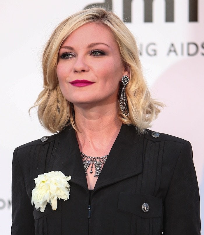 A closer look highlights her luminous beauty styling, with softly waved blonde hair, berry-toned lips, and ornate drop earrings that frame her face with vintage glamour