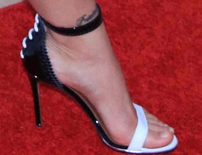 A close-up reveals her Givenchy “Nadia” sandals with a white toe strap, glossy black ankle strap, and slim stiletto heel framing her tattooed foot