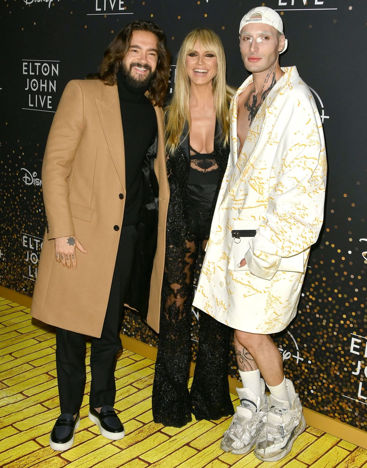 Tom Kaulitz, Yannik Zamboni, and Heidi Klum pose together at Elton John’s Farewell Yellow Brick Road Dodger Stadium event, with Klum standing out in a sheer black lace Moschino suit paired with sleek pointed heels, flanked by Kaulitz’s tailored camel coat and Zamboni’s avant-garde white ensemble