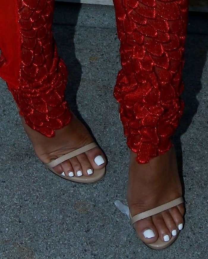 Christina Milian shows off her white pedicure in nude Stuart Weitzman “Nudistsong” sandals, adding a subtle contrast to her bold red lace trousers