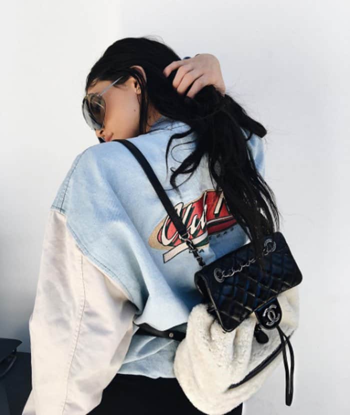 Kylie Jenner serves retro-cool vibes in a faded Coca-Cola varsity jacket, accessorizing with oversized sunnies and her signature Chanel shearling mini backpack