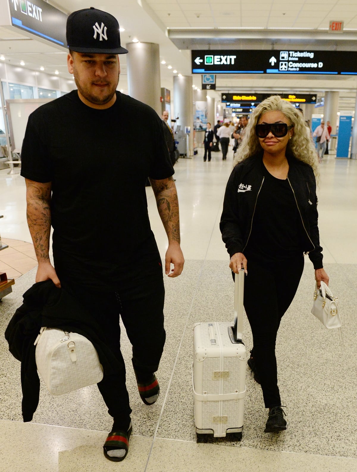 Rob Kardashian towers beside Blac Chyna as the couple arrives at the airport in coordinated monochrome travel looks, with Chyna styling oversized sunglasses, black sneakers, and a compact white handbag